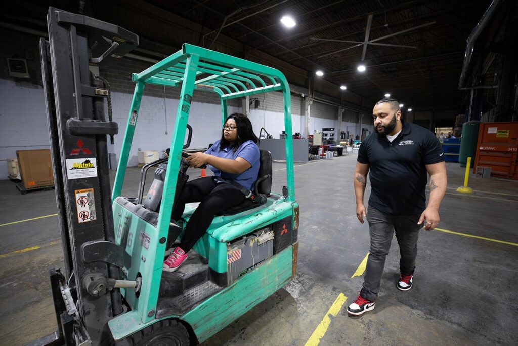 VersAbility Tech instructor walking alongside a student operating a forklift in a warehouse.