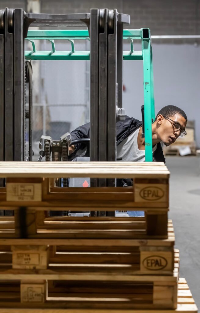 VersAbility Tech student operating a forklift to align forks with a stack of wooden pallets.
