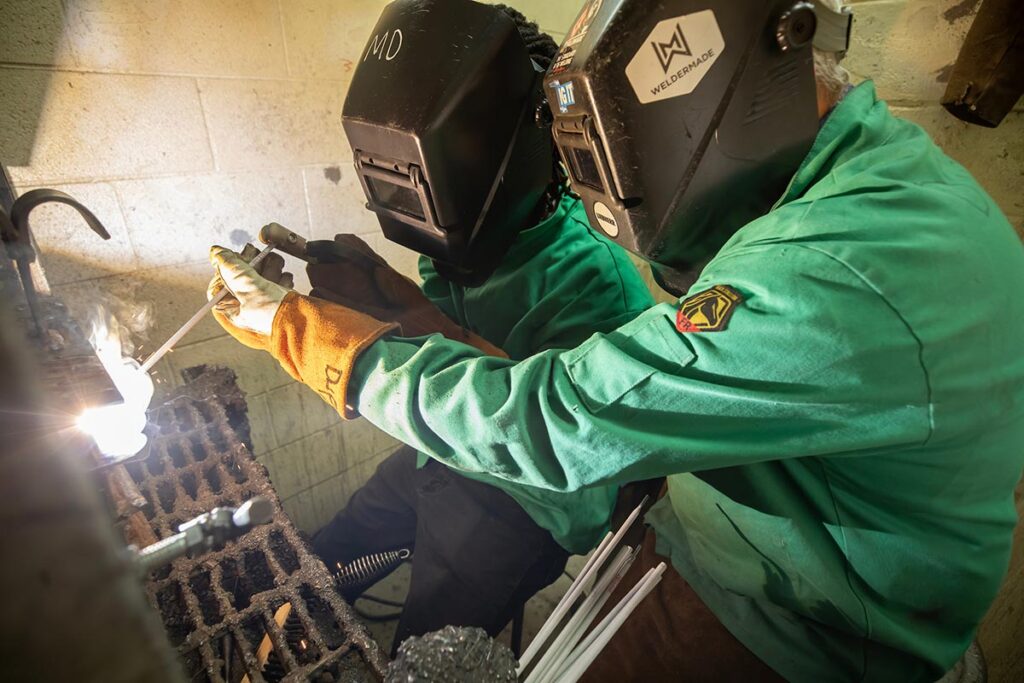 Side view of two VersAbility Tech students in full welding gear practicing at a welding station.
