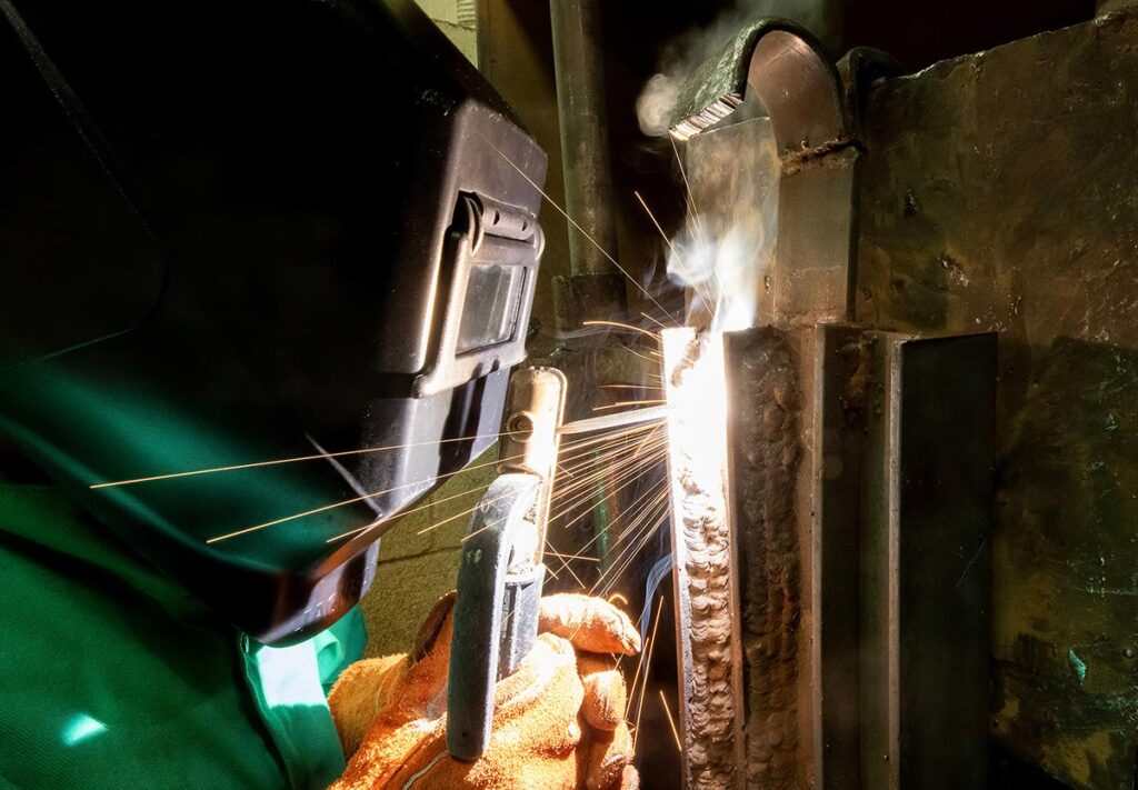 Close-up of a VersAbility Tech student welding along a vertical seam with sparks and smoke.