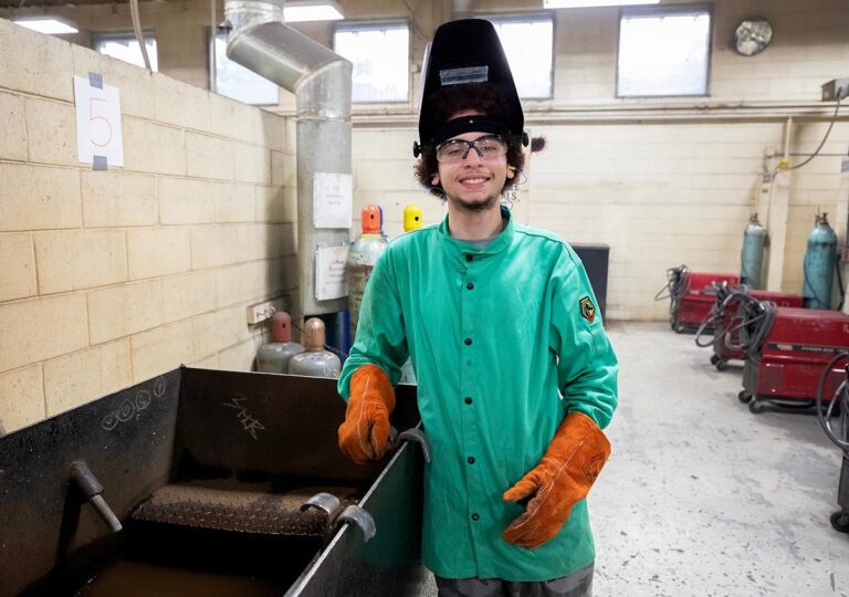 VersAbility TECH welding student smiling in a green jacket and helmet in the welding shop.