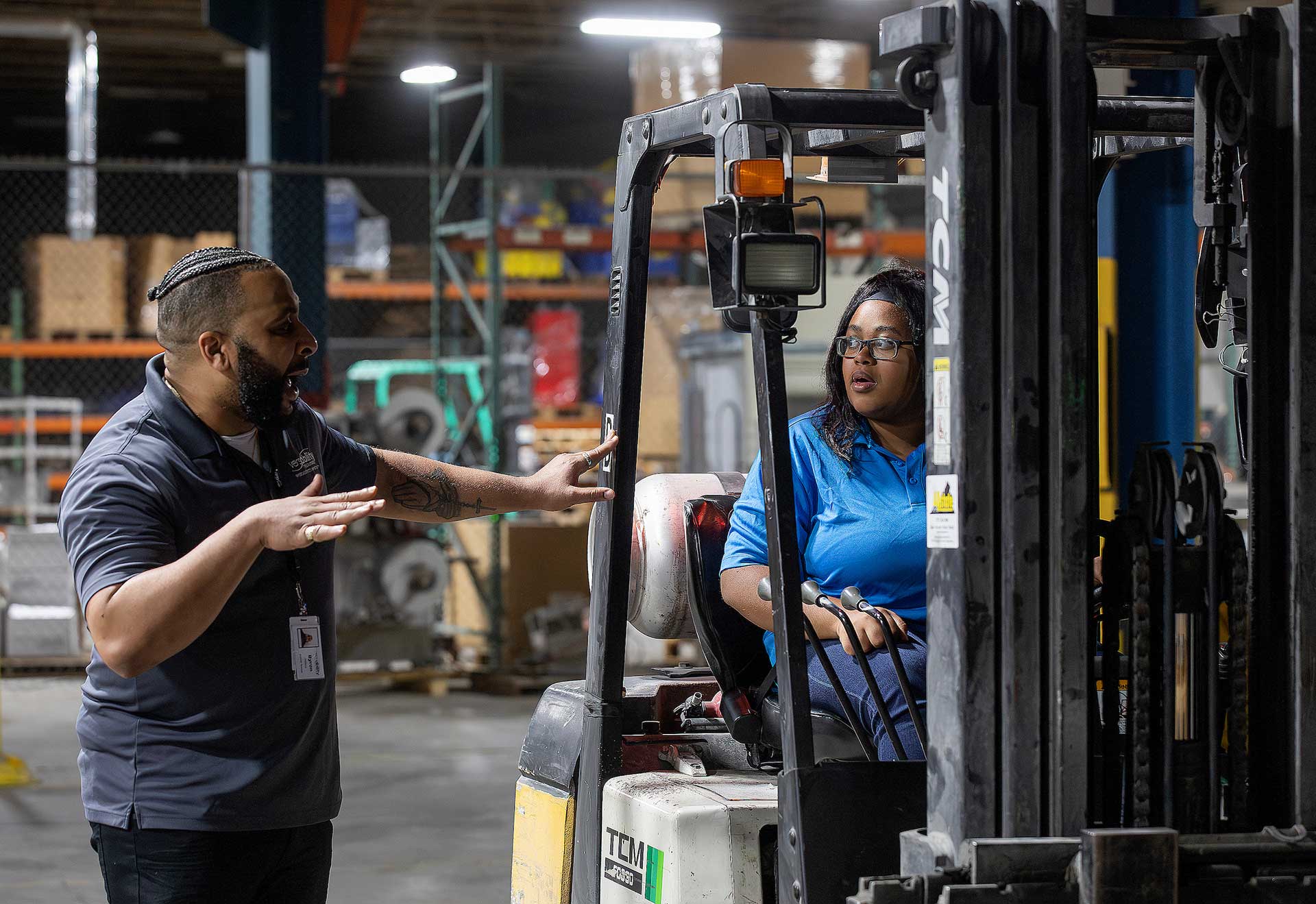 Instructor gesturing and explaining forklift controls to a student seated in the operator's chair.